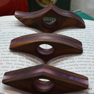 May include: Three wooden page holders with a central circular cutout and a translucent purple resin inlay. Each holder features a curved, elongated diamond shape with a smooth finish, designed to rest on the open pages of a book.
