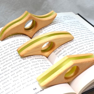 May include: Three wooden page holders with a central circular opening and a light green resin inlay running along the top edge. Each holder features a curved, elongated shape designed to rest on the open pages of a book for hands-free reading.