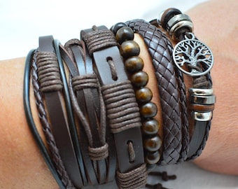 May include: A multi-layered bracelet set featuring dark brown leather straps, braided leather cords, and a row of smooth, dark brown wooden beads. The set includes a silver-toned metal charm depicting a tree of life, accented with small silver-toned metal rings.
