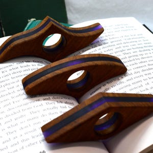 May include: Three wooden page holders with a central circular cutout and a translucent purple resin inlay running horizontally through the center of each piece. The page holders are shaped with pointed ends and a curved profile for holding books open.