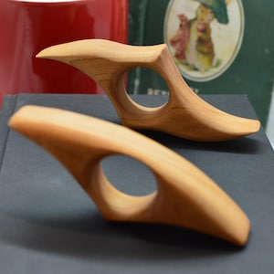 May include: Two hand-carved wooden objects with a smooth, light brown finish. Each piece features a curved, elongated shape with a circular hole in the center, resembling a stylized handle or grip, resting on a dark surface.