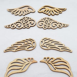 May include: Six pairs of wooden angel wings in various designs. The wings are cut from light-colored wood and arranged on a white surface. The wings feature detailed cut-out patterns, perfect for crafts or decorations.