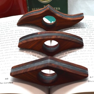 May include: Three wooden page holders with a dark brown grain and a central gray inlay. Each holder features a smooth, curved, elongated shape with a circular hole in the middle, designed to rest on the open pages of a book for hands-free reading.