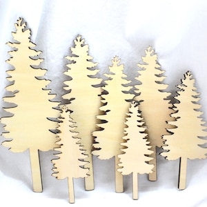 May include: A collection of six light-coloured wooden tree cutouts in varying heights. Each piece features a detailed, jagged silhouette representing a pine tree with a straight trunk base, crafted from smooth, unfinished light beige wood material.