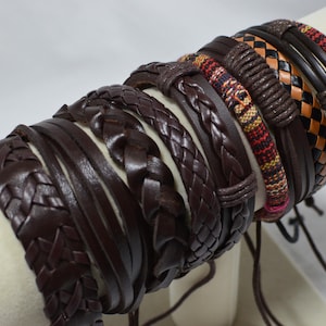 Puede incluir: Una colección de múltiples pulseras marrones con varios diseños, incluyendo hebras trenzadas tipo cuero, bandas de cordón liso y patrones de tela tejida con acentos en rojo, amarillo y negro, exhibidas juntas en un soporte cilíndrico.
