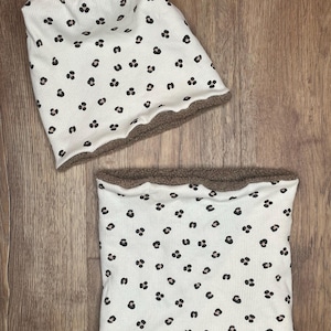 May include: A white beanie and neck gaiter set with a black leopard print pattern. The beanie and neck gaiter have a brown, fuzzy lining. The set is made of a ribbed fabric.