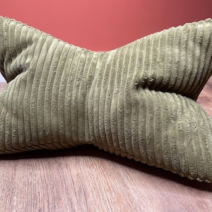 May include: An olive green corduroy pillow with a distinctive shape, lying on a wooden floor. The pillow features a ribbed texture and a small tag. The background is a red wall.