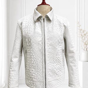 Puede incluir: Una chaqueta blanca con un patrón de textura de piel de cocodrilo en todo el exterior. La prenda incluye un cuello clásico, un cierre de cremallera metálica vertical en toda la longitud y dos bolsillos laterales con cremallera en el frente.