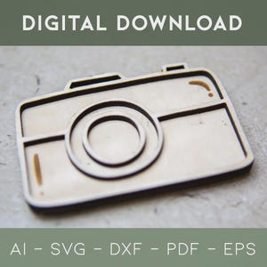 May include: A wooden camera-shaped cutout with a detailed design, featuring a lens and rectangular body. The text "DIGITAL DOWNLOAD" is at the top, and "AI - SVG - DXF - PDF - EPS" is at the bottom. The cutout is light brown.