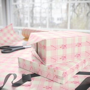 May include: Two rectangular gift boxes wrapped in pink and white gingham paper with candy cane illustrations. A roll of matching wrapping paper, scissors, and black ribbon are also visible.