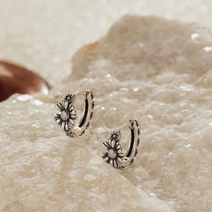 May include: A pair of silver hoop earrings featuring a detailed, raised sunflower or daisy floral motif along the outer edge. The metal has an oxidized finish that highlights the intricate petal and center textures of the flower design.