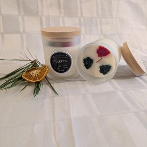 Könnte beinhalten: Zwei Kerzen aus mattiertem Glas mit Holzdeckeln, eine aufrecht und eine auf der Seite. Die aufrechte Kerze hat ein Etikett mit der Aufschrift "Yuletide". Die Kerzen sind mit getrockneten Pflanzen dekoriert. Ein Kiefernzweig und eine getrocknete Orangenscheibe liegen neben den Kerzen.