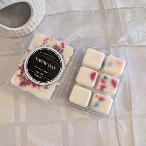 Könnte beinhalten: Zwei transparente Kunststoffbehälter mit Sojawachs-Schmelzen. Ein Behälter trägt die Aufschrift "Snow Day" und ist mit roten und grünen Streuseln verziert. Der andere Behälter hat sechs quadratische Wachs-Schmelzen mit roten und grünen Akzenten.