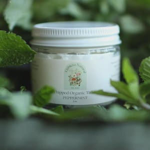 Può includere: Un barattolo di vetro con coperchio ed etichetta bianchi, circondato da foglie verdi. L'etichetta recita "Whipped Organic Tallow PEPPERMINT" con un disegno floreale. Il barattolo contiene una sostanza bianca, possibilmente un prodotto per la cura della pelle.