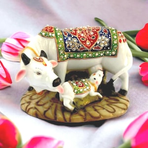 May include: A white ceramic figurine of a cow licking its calf. The cow wears a decorative blanket with red, blue, and green patterns accented with small clear rhinestones. The calf also wears a matching green blanket. Both stand on a tan base.