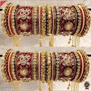 May include: Two sets of ornate, red and gold bangles. Each set features intricate floral and sunburst patterns, embellished with pearls and sparkling stones. Gold chains hang from the bangles, adding a touch of elegance. The bangles are likely for a special occasion.
