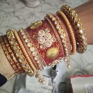 May include: A collection of gold and red bangles and bracelets. The bangles feature intricate designs with clear gemstones and gold accents. Some bangles have a pearl-like appearance. The bracelets are stacked on top of each other.