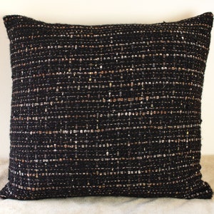 May include: Black throw pillow with a textured woven fabric. The fabric has a subtle shimmer and a subtle pattern of small white and brown threads.