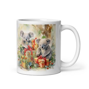 May include: White ceramic mug featuring a watercolor illustration of two koalas with gifts. The koalas are surrounded by presents with red bows and greenery. The mug has a white handle and a glossy finish.