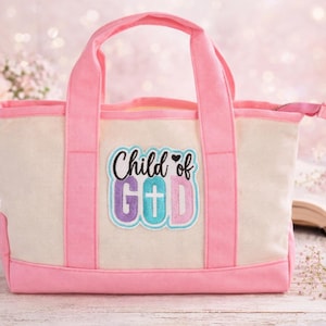 May include: A pink and cream-colored tote bag with pink handles and trim. The bag features the text "Child of God" in a colorful patch with a cross. The bag is made of canvas material.