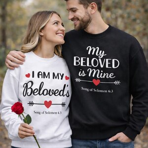May include: A couple wearing matching sweatshirts. One sweatshirt is white with the text "I AM MY Beloved's" and the other is black with the text "My BELOVED is Mine". Both sweatshirts have a red heart and the text "Song of Solomon 6:3".