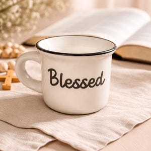 May include: White ceramic mug with a black rim and handle, featuring the word "Blessed" in black script. The mug sits on a beige linen napkin, with a blurred background including a book and flowers. A simple, elegant design.
