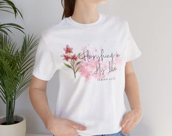 May include: White t-shirt featuring a floral design with pink and red flowers and the text "Flourishing in His Love" and "Isaiah 61:11". The shirt has short sleeves and a crew neck.