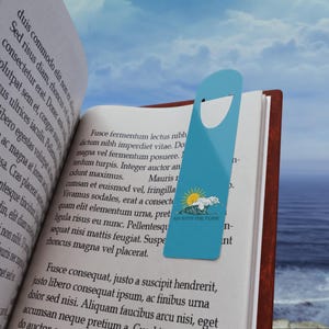 May include: A light blue bookmark with a cutout at the top is placed in an open book. The bookmark features a sun and wave graphic with the text "GO WITH THE FLOW". The book has brown edges and is open to a page of text.