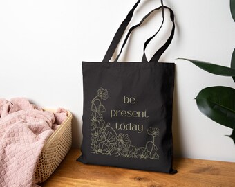 May include: Black tote bag with the text "be present today" in a serif font. The bag features a floral design in the lower left corner and is made of a sturdy fabric. The handles are black.