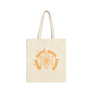 May include: Cream-colored tote bag with a floral design and the words "quiet mind open heart" in orange. The bag has two handles and is made of a canvas-like material. The floral design is in the center of the bag.