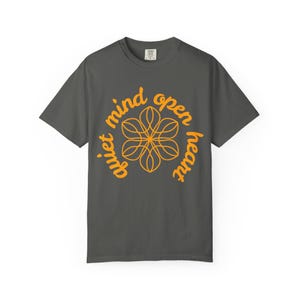 May include: A charcoal gray t-shirt with the words "quiet mind open heart" in orange script, encircling an orange floral design. The short-sleeved shirt is made of a soft material and is suitable for casual wear.
