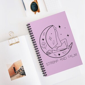 May include: A spiral-bound notebook with a light purple cover featuring a black line drawing of a person in a yoga pose inside a crescent moon. The words "STRONG AND CALM" are printed below the illustration.