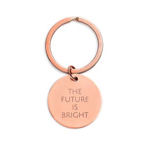 May include: Rose gold keychain with a circular pendant. The pendant has the engraved text "THE FUTURE IS BRIGHT". The keychain includes a split ring and a smaller ring connecting the pendant. The metal has a shiny finish.