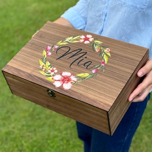 Op de afbeelding: Een rechthoekige houten aandenkendoos met een donkerbruine houtnerfafwerking. Het deksel is voorzien van een bedrukte bloemenkrans in rood-, roze- en groentinten, rondom de naam Mia geschreven in een zwart cursief lettertype. Een metalen sluiting is zichtbaar.