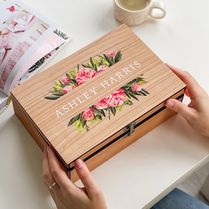 Puede incluir: Una caja de madera con un diseño floral y el nombre "ASHLEY HARRIS" impreso en la tapa. La caja tiene un cierre de broche y un borde negro. La caja está sobre una superficie blanca. También se ve una taza de café.
