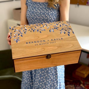 Pode incluir: Uma caixa de madeira com tampa articulada, decorada com desenhos florais azuis e dourados. A caixa é personalizada com os nomes "BRANDON + KAYLA" e a data "20 MARÇO 2027". Possui um fecho de metal.