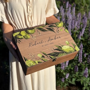 May include: A rectangular wooden keepsake box featuring a printed design of green leaves and small white flowers. The lid is personalised with the text "Robert + Amber" and the date "18 JULY 2026" in black script and sans-serif fonts.