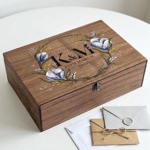 Op de afbeelding: Een rechthoekige houten aandenkendoos met een metalen sluiting, voorzien van een bedrukte bloemenkrans in blauwe en groene tinten rondom de zwarte tekst K&M en 25TH MARCH 2027. Voor de doos liggen drie enveloppen, waaronder één met een lakzegel.