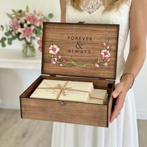 Op de afbeelding: Een rechthoekige houten bewaardoos met een scharnierend deksel voorzien van de tekst "FOREVER & ALWAYS" en bloemillustraties. De open doos bevat een stapel crèmekleurige enveloppen die met een dun lint zijn samengebonden.