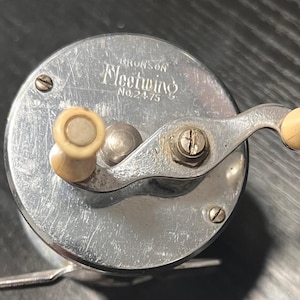 May include: Vintage Bronson Fleetwing No. 24-75 fishing reel. The reel is silver-toned with a cream-colored handle and knobs. The name "Fleetwing" is printed on the reel. A classic piece of fishing equipment.