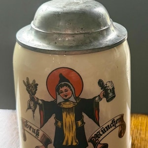 May include: A cream-colored ceramic beer stein with a metal lid and a decorative thumb lift. The front features a colorful illustration of the Münchner Kindl holding a radish and a beer mug, with the text "Gruß aus München" on a banner.
