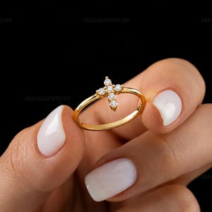 Pode incluir: Um anel fino de ouro com um pequeno motivo em forma de cruz adornado com cinco pedras claras de corte redondo. A banda é lisa e polida, apresentando um design simples com o elemento de cruz centrado no topo do anel circular.