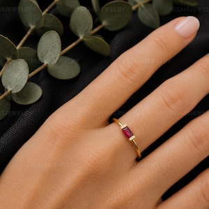 May include: A thin gold ring featuring a single rectangular baguette-cut ruby gemstone set horizontally. The ruby is flanked by two tiny, clear round accent stones on each side, creating a delicate and refined piece of jewelry.