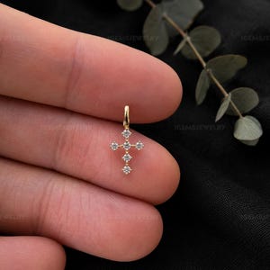 May include: A small gold cross pendant featuring six clear, round-cut stones set in a vertical and horizontal arrangement. The pendant includes a small gold bail at the top for attachment to a chain, showcasing a delicate and refined metal design.