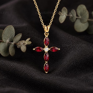 Puede incluir: Un collar de cadena dorada con un colgante en forma de cruz. El colgante está compuesto por cinco piedras preciosas de color rojo intenso con talla marquesa dispuestas en cruz, con una piedra transparente redonda engastada en el centro.