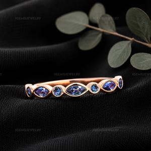 May include: A rose gold band ring featuring an alternating pattern of marquise-cut and round-cut blue-violet stones set in a bezel style. The ring displays a delicate metal construction with a polished finish, showcasing a symmetrical arrangement.