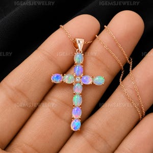 May include: A rose gold cross pendant necklace featuring nine oval-shaped cabochon opals with iridescent blue and green flashes, centered with a small clear round gemstone, suspended on a delicate rose gold chain with a matching metal bail.