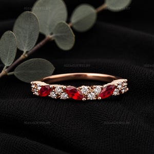 May include: A rose gold band ring featuring three marquise-cut red gemstones alternating with clusters of small, round, clear white stones. The ring has a delicate, symmetrical design with a polished metal finish and a thin, circular band structure.
