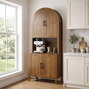 May include: A tall, arched wooden hutch with vertical slat detailing and black hardware. The unit features two upper doors, an open middle shelf containing a silver espresso machine and stacked mugs, and two lower doors with four short legs.