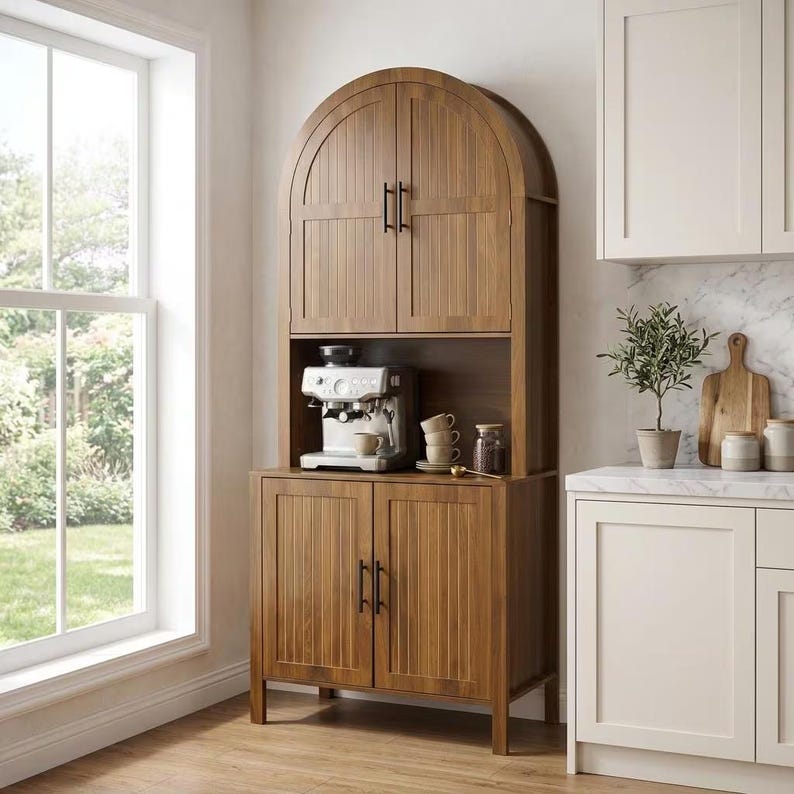 May include: A tall, arched wooden hutch with vertical slat detailing and black hardware. The unit features two upper doors, an open middle shelf containing a silver espresso machine and stacked mugs, and two lower doors with four short legs.